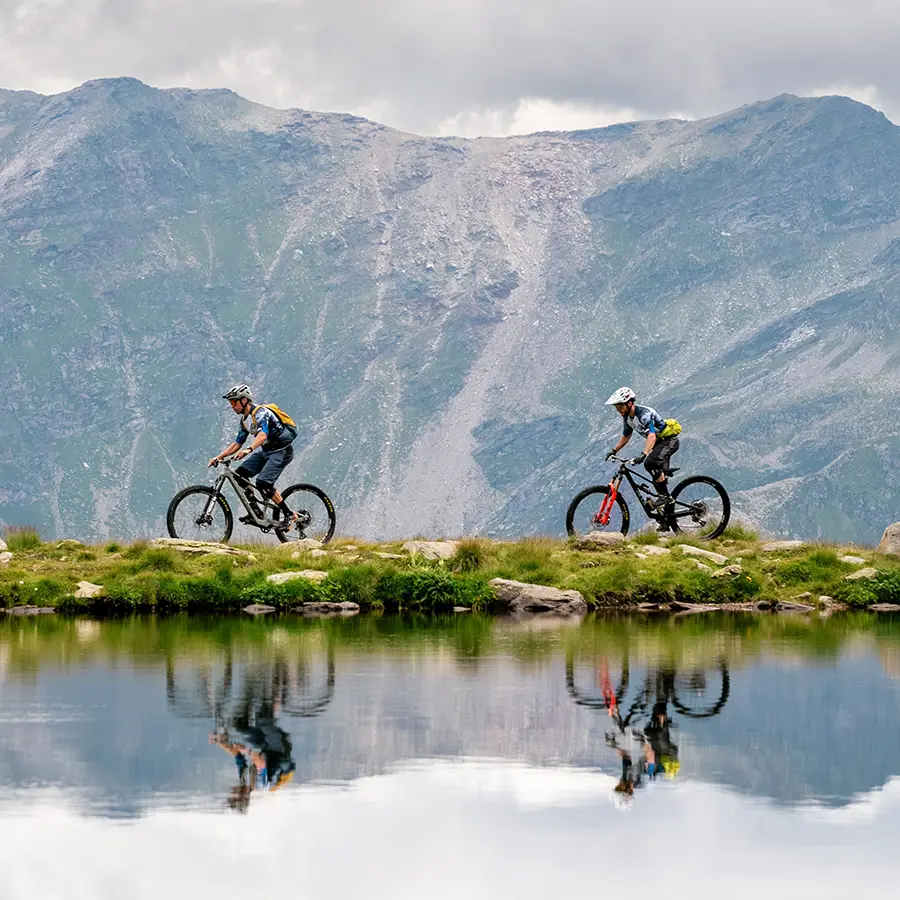 Two mountain bikers riding along the lake's edge, surrounded by scenic mountains and reflecting water.